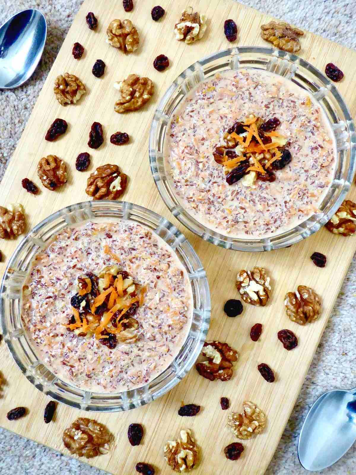 2 carrot cake flaxseed pudding servings in bowls on a chopping board surrounded by raisins, walnuts and 2 spoons.